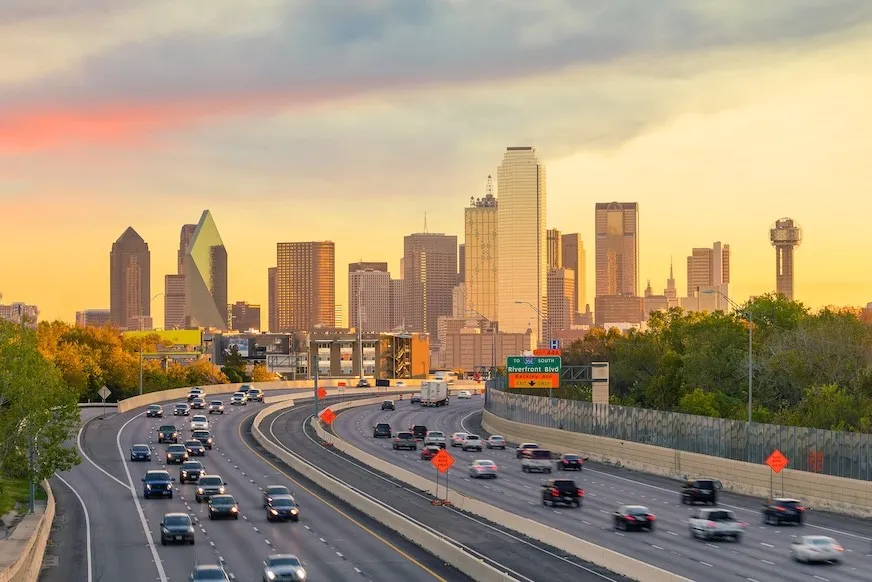 Traffic on a Dallas freeway at sunset, with the downtown Dallas skyline ahead